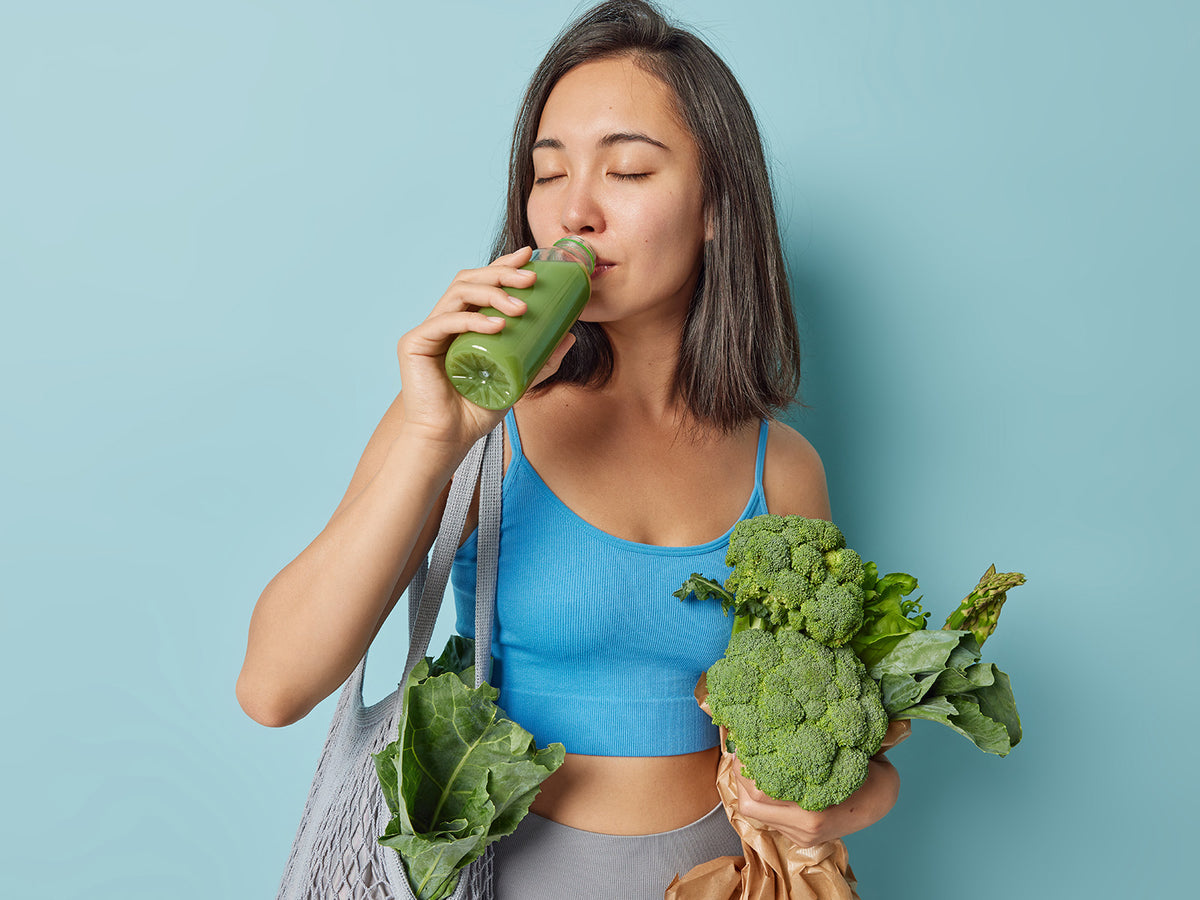Woman in athletic wear holding fresh leafy greens and drinking green juice, symbolising a clean and nutrient-rich lifestyle.